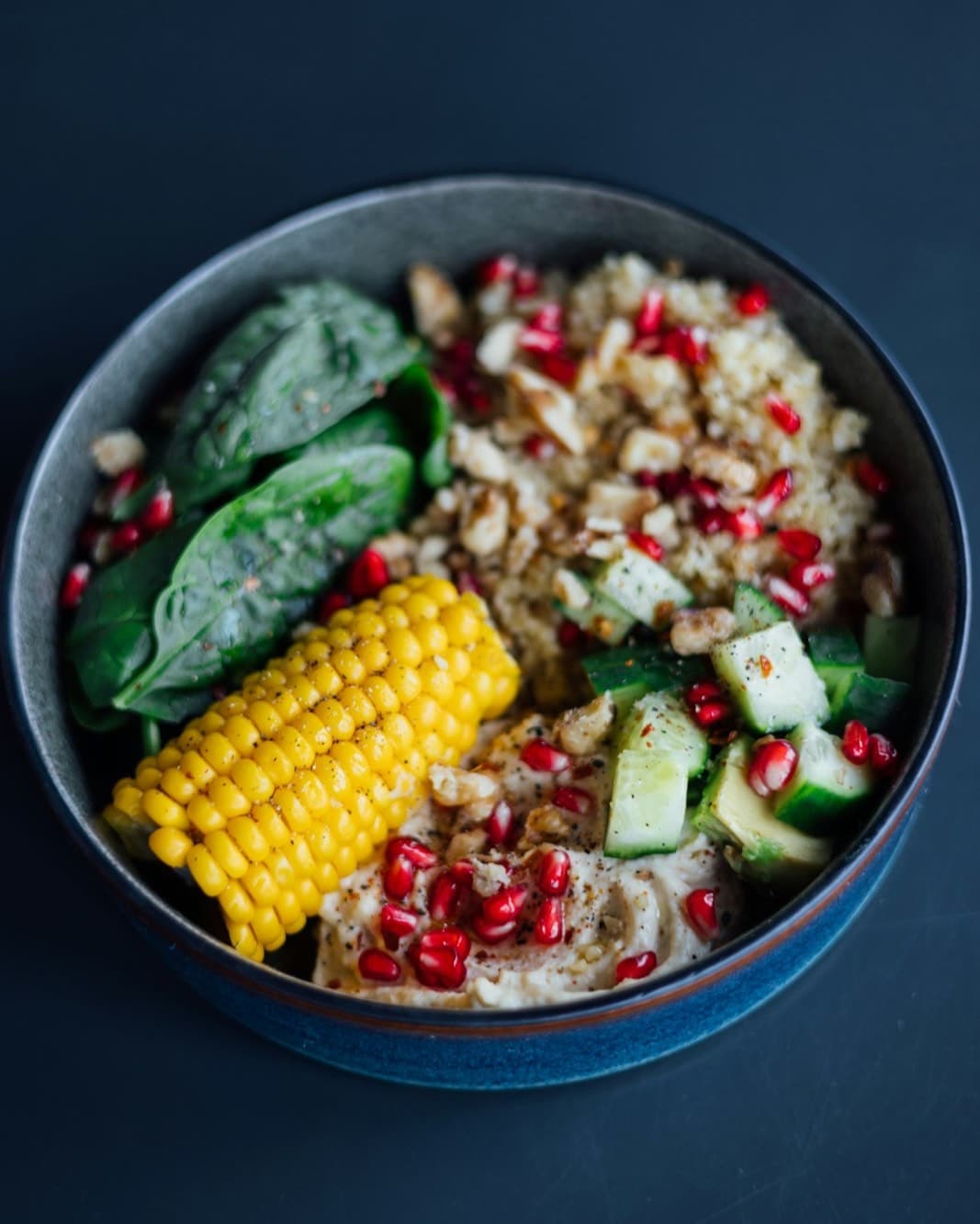 Overhead view of a grain bowl with a corn cob, spinach, cucumber and pomegranate seeds on a dark table.