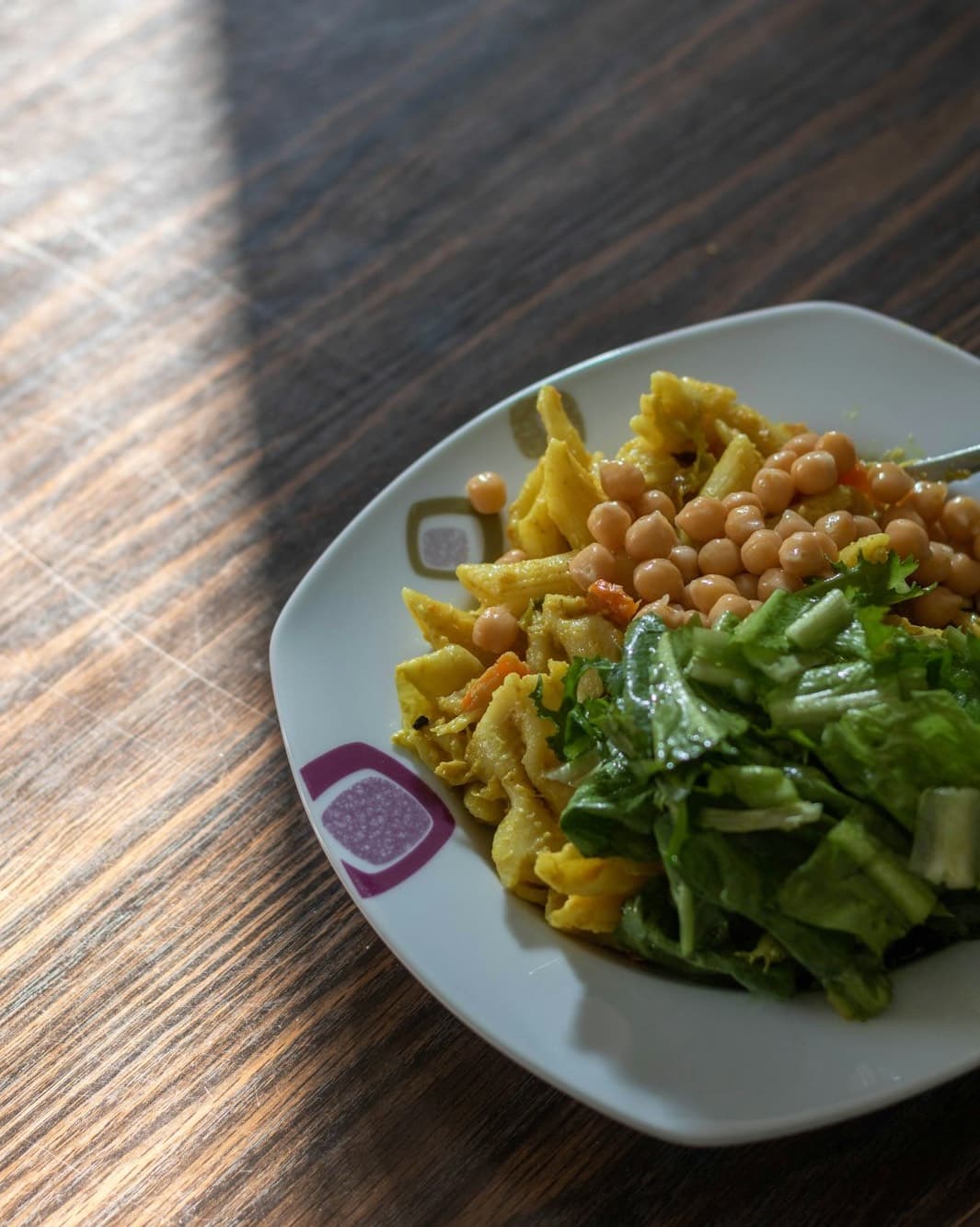 A plate of short pasta tossed with chickpeas next to green salad, in warm window light.