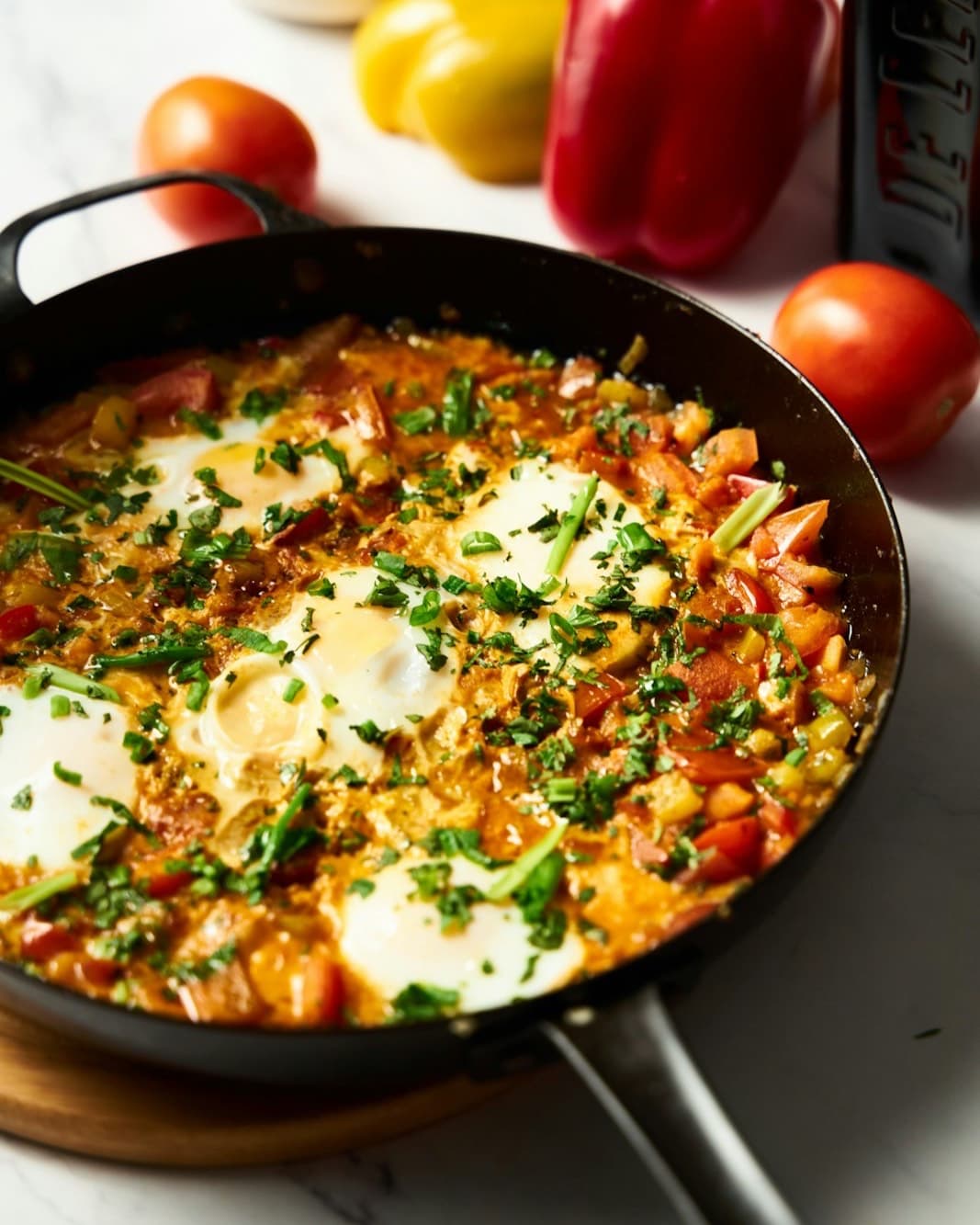 A skillet of shakshuka, eggs set in a red pepper and tomato sauce with herbs, beside fresh peppers.