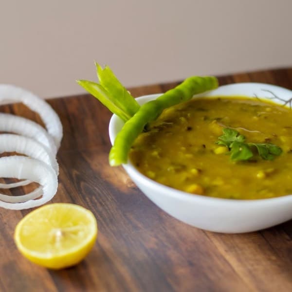 A white bowl of yellow lentil dal with a green chilli, onion rings and half a lemon on a wooden table.