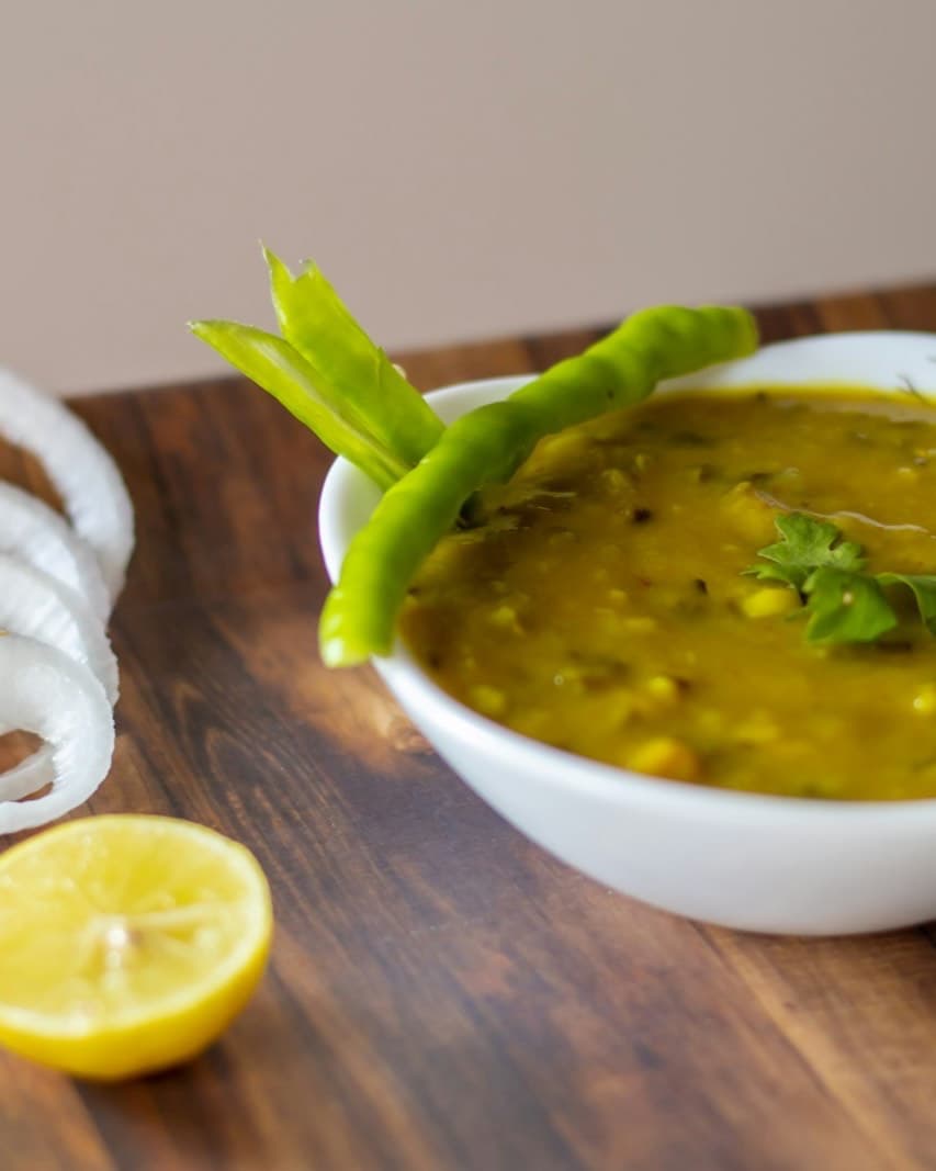 A white bowl of yellow lentil dal with a green chilli, onion rings and half a lemon on a wooden table.