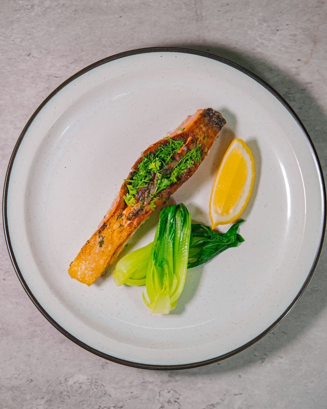 A seared salmon fillet with herbs, steamed bok choy and a lemon wedge on a white plate.