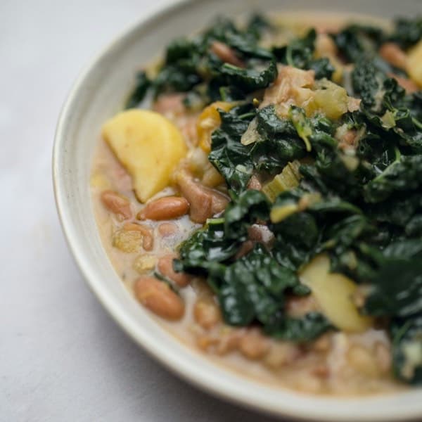 A bowl of bean soup with dark cavolo nero kale and potato on a pale table.